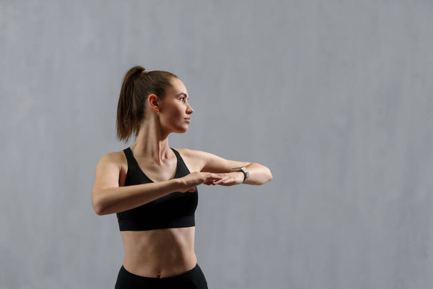 A young sporty woman in sports clothes stretching arms against grey background, copy space - Photo, Image