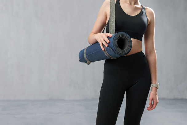 A sports woman in fashion black sport clothes carrying fitness mat in gym, over gray background - Foto, Imagem