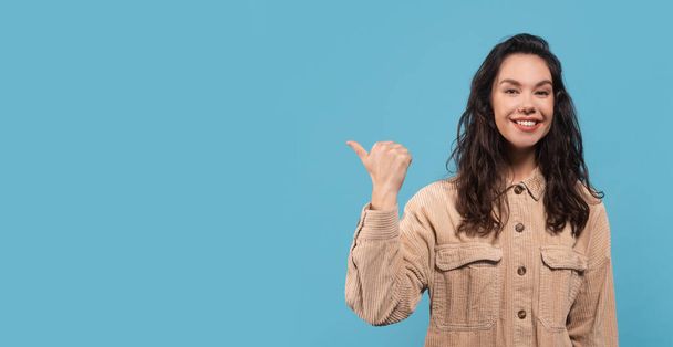 Muotokuva iloinen nuori valkoihoinen nainen brunette opiskelija rento osoittaa sormen tyhjään tilaan, eristetty sinisellä pohjalla, studio, panoraama. Hyvä mainos ja tarjous, kasvojen ilme, myynti ja neuvonta - Valokuva, kuva