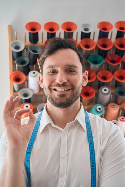 Πορτρέτο του χαμογελαστού νεαρού άνδρα ράφτη δείχνει ok χειρονομία, ενώ στέκεται μπροστά από τη θήκη με νήμα - Φωτογραφία, εικόνα