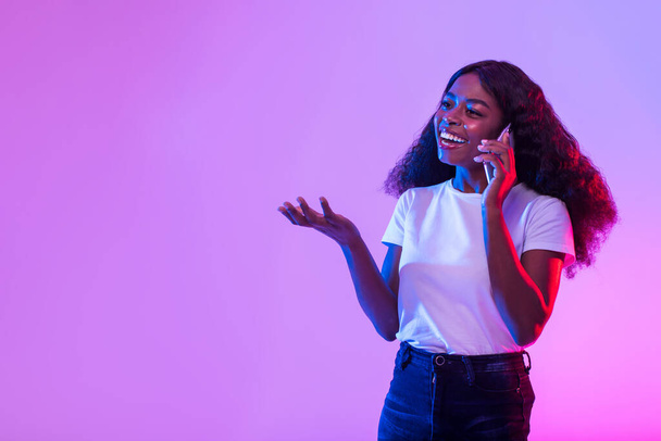 Happy millennial black woman talking on smartphone in neon light, free space. Positive young African American female making phone call, smiling and having friendy conversation - Fotografie, Obrázek