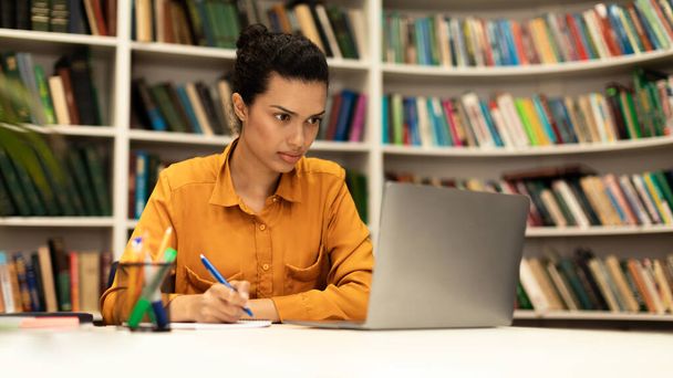 Зосереджена жінка використовує ноутбук і пише в блокноті, вивчає онлайн в інтер'єрі бібліотеки, переглядає Інтернет, переглядає вебінар або лекцію, панораму з вільним простором
 - Фото, зображення