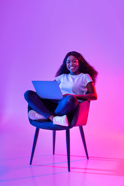 Pełna długość czarującej młodej czarnej kobiety pracującej online, siedzącej w fotelu i korzystającej z laptopa w neonowym świetle. Pozytywne millenium pani przeglądanie Internetu, oglądanie webinarium na komputerze przenośnym - Zdjęcie, obraz