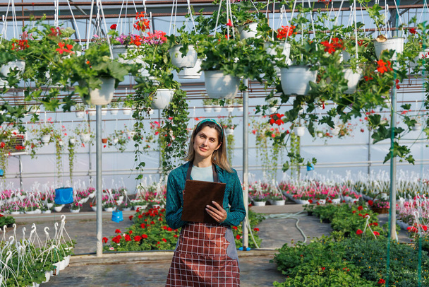 カメラのノートを持っている女性の庭師は温室でノートを作る。多くの花を持つ大規模な工業用温室で作業員は植物を調べ - 写真・画像