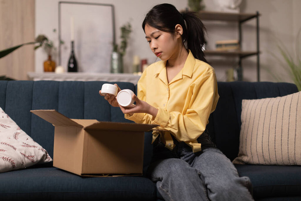 Asya 'lı genç bir kadın karton kutuları açıyor. Kozmetik kavanozları tutuyor. Evdeki koltukta oturan güzellik ürünleri alıyor. Kozmetik ve Özbakım, Teslimat Hizmeti Konsepti - Fotoğraf, Görsel
