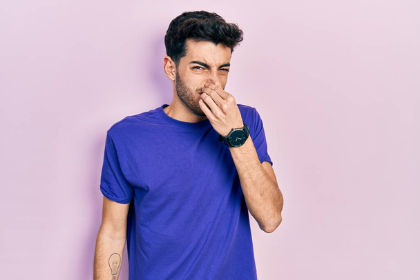 Junger hispanischer Mann in lässigem T-Shirt, der etwas Stinkendes und ekelhaften, unerträglichen Geruch riecht und mit den Fingern auf der Nase den Atem anhält. schlechter Geruch  - Foto, Bild