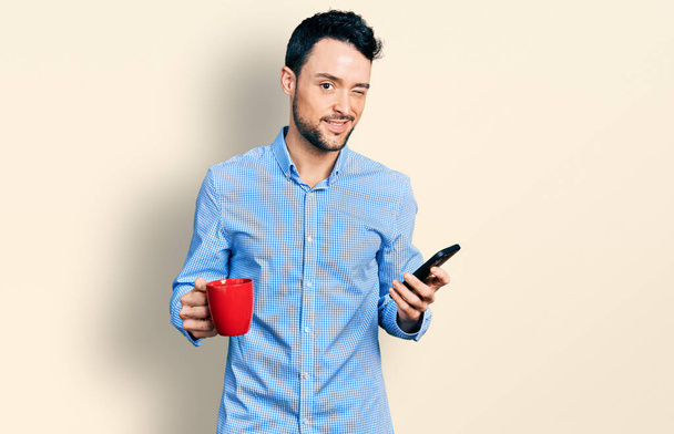 Латиноамериканец с бородой с помощью смартфона и выпив чашку кофе подмигивая глядя в камеру с сексуальным выражением лица, веселым и счастливым лицом.  - Фото, изображение