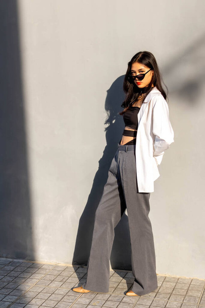 Concept à la mode, Mode femme à poser en lunettes de soleil et pantalon avec chemise blanche près du mur gris. - Photo, image
