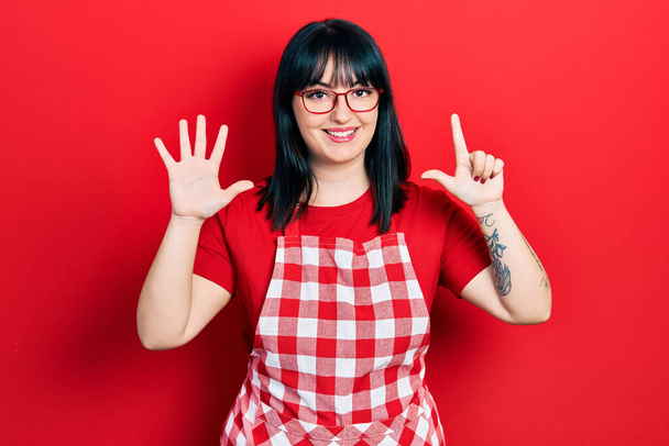 Jonge Spaanse vrouw draagt een keukenschort en een bril met vingers nummer zeven en glimlacht zelfverzekerd en gelukkig.  - Foto, afbeelding
