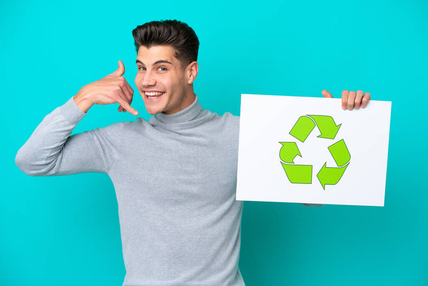Young handsome caucasian man isolated on blue bakcground holding a placard with recycle icon and doing phone gesture - Foto, Imagen