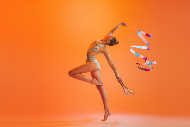 Dance, emotions. Young sportive girl, rhythmic gymnastics artist dancing isolated on orange color background. Concept of sport, action, aspiration, education, active lifestyle - Photo, Image