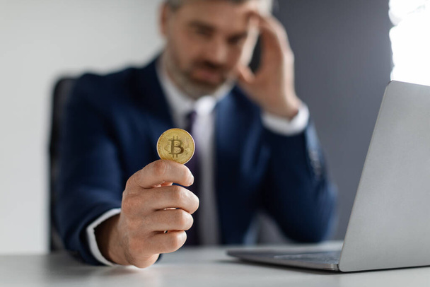 Crypto Failure. Upset Businessman Holding Bitcoin While Sitting At Desk With Laptop In Office, Depressed Male Entrepreneur In Suit Touching Head In Despair, Losing Money On Online Trading - Photo, Image