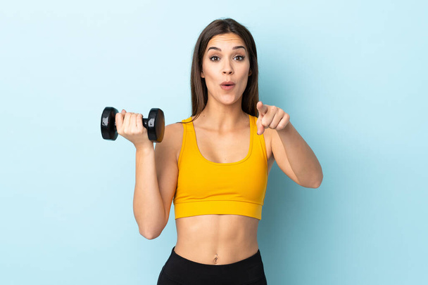 Junge kaukasische Frau macht Gewichtheben isoliert auf blauem Hintergrund überrascht und zeigt nach vorne - Foto, Bild