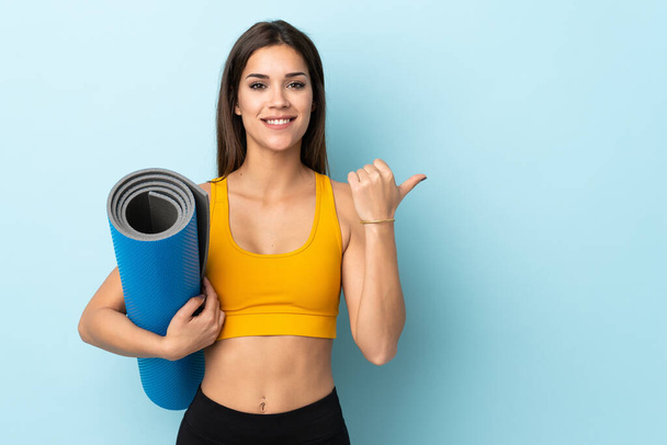 Young sport woman with mat isolated on blue background pointing to the side to present a product - Fotó, kép