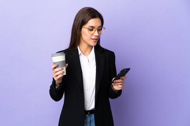 Mor arka planda izole edilmiş bir iş kadını elinde kahve ve bir cep telefonu tutuyor. - Fotoğraf, Görsel