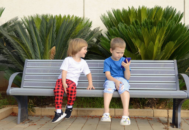 Dos niños pequeños están sentados en un banco en el parque y jugando con un smartphone. Niños y artilugios. Los hermanos están hablando por teléfono, viendo dibujos animados. - Foto, imagen