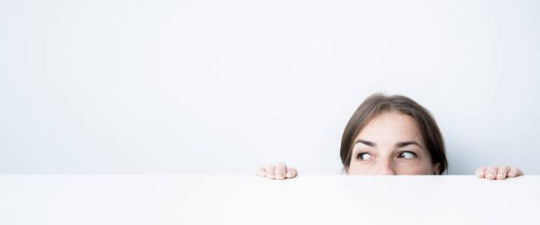 Νεαρή γυναίκα κοιτάζει στο πλάι κοιτάζοντας έξω κάτω από ένα λευκό τραπέζι ενάντια σε ένα λευκό τοίχο. Μπάνερ. - Φωτογραφία, εικόνα