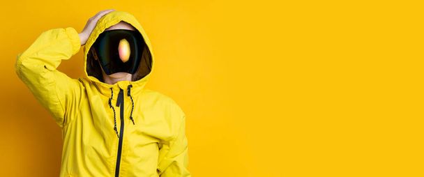 A young woman in a mask of cyberpunk goggles in a yellow jacket holds her head with her hand on a yellow background. Banner. - Photo, Image