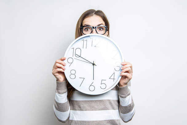 Young woman in glasses peeks out from under the wall clock in surprise on a light background  - Photo, Image