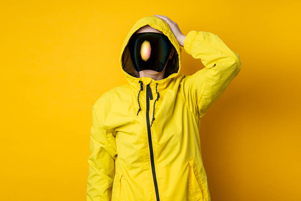 Młoda kobieta w masce gogli cyberpunkowych w żółtej kurtce trzyma głowę na żółtym tle.. - Zdjęcie, obraz