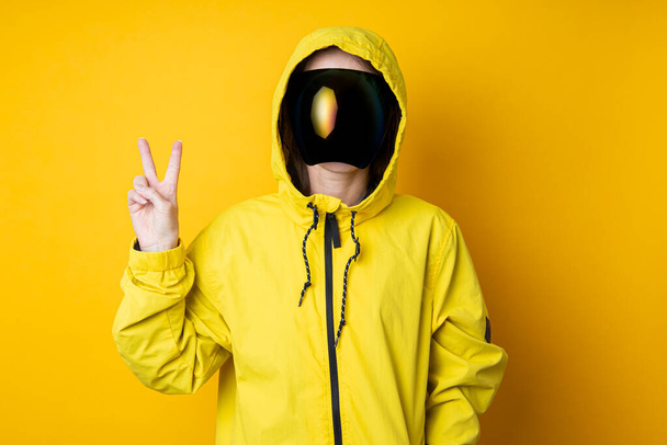 Mladá žena v masce kyberpunkových brýlí ve žluté bundě ukazuje mírové vítězství na žlutém pozadí. - Fotografie, Obrázek