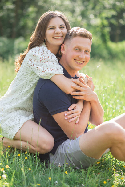 Affectionate young couple sitting on grass in park, looking at camera. Young man and woman enjoying summer day. Happy people in love, honey moon.  - Foto, imagen
