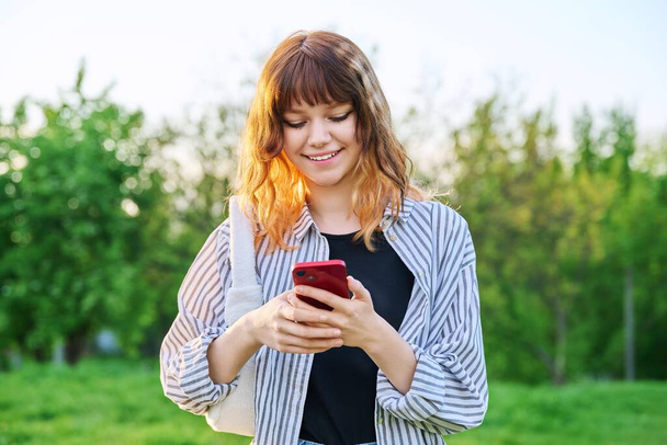 Outdoor portrait of trendy young red-haired girl with smartphone in hand, green trees sky background. Smiling teenage female student looking at phone. Youth, beauty fashion, lifestyle, summer concept - Zdjęcie, obraz