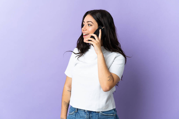 Junge kaukasische Frau isoliert auf lila Hintergrund hält ein Gespräch mit dem Handy mit jemandem - Foto, Bild