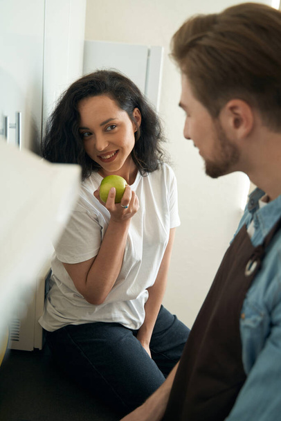 Woman with green apple in hand sitting on kitchen counter and smiling at man in apron - Foto, Imagem