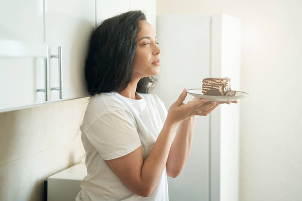 Woman seated on kitchen counter smelling fancy cake on plate in her hands - Fotó, kép