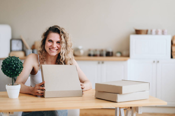 Young Cheerful Girl Opens Flat Cardboard Box Sitting At The Wooden Table. Girl Got Pizza Delivery - Foto, Imagen