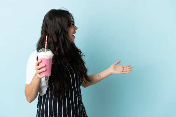 jonge vrouw met aardbei milkshake geïsoleerd op blauwe achtergrond met verrassing uitdrukking terwijl op zoek kant - Foto, afbeelding