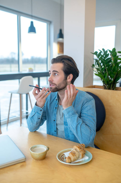 Voice message. Young adult man looking away talking holding smartphone at mouth level sitting at table in cafe during daytime - Фото, изображение