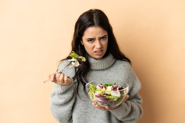 Young caucasian woman isolated on beige background holding a bowl of salad with sad expression - Foto, imagen