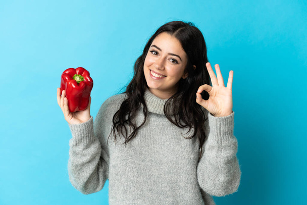 Young caucasian woman holding a pepper isolated on blue background showing ok sign with fingers - Photo, Image
