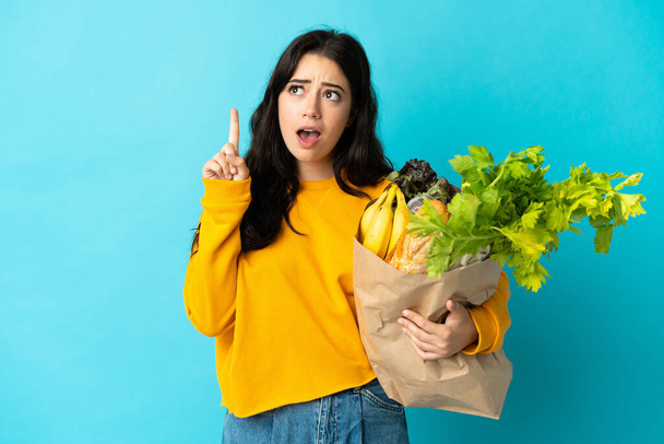 Fiatal nő kezében egy élelmiszer bevásárló táska elszigetelt kék háttér gondolkodás egy ötlet rámutatva az ujját fel - Fotó, kép