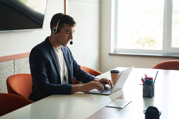 Человек говорит в микрофон гарнитуры во время ввода на ноутбуке клавиатуры в офисе во время онлайн-работы - Фото, изображение