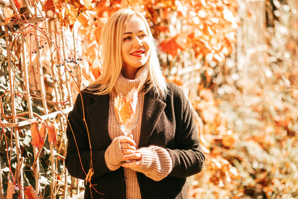 Beautiful blonde 35 years old in black coat and beige sweater holds glass with autumn cocktail in her hands and enjoys sunny day in city park. Colors of autumn. Seasons. Rest and pleasure. Leaf fall. - Фото, изображение