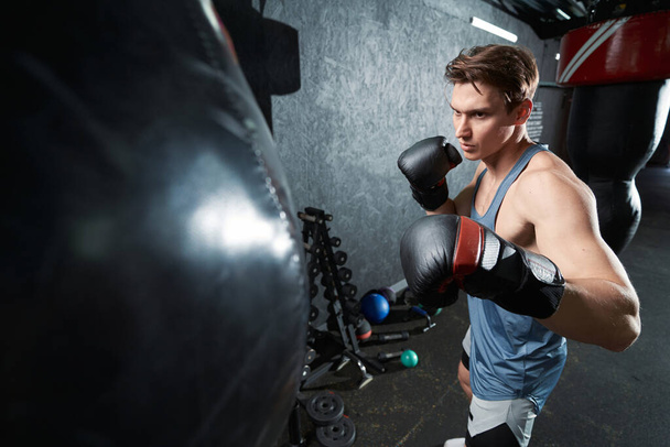 Serious concentrated young muscular man in boxing gloves standing before punching bag - Foto, afbeelding