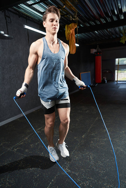 Full-length πορτρέτο του ταιριάζει νεαρός πηδώντας σχοινί κατά τη διάρκεια προπόνηση γυμναστήριο - Φωτογραφία, εικόνα