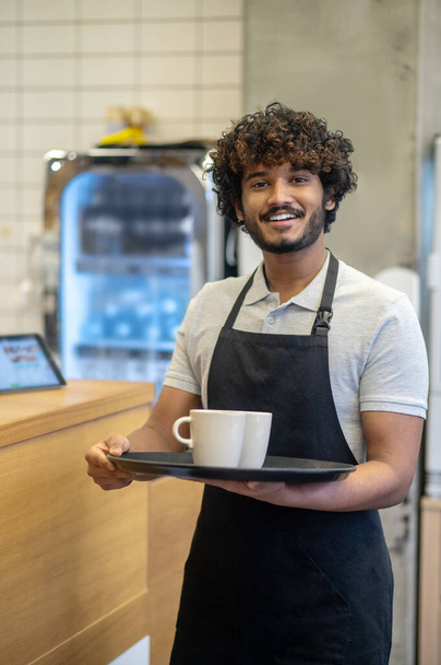 Lucky day. Young adult indian man in apron holding cups on tray smiling confidently at camera standing in cafe kitchen - Foto, imagen