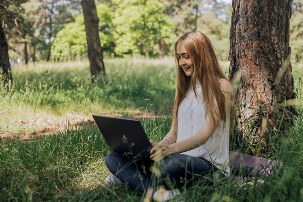 On the banner, a young girl works with a laptop in the fresh air in the park, sitting on the lawn. The concept of remote work. Work as a freelancer. The girl takes courses on a laptop and smiles - Photo, Image