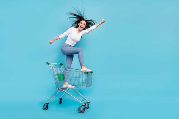 Full size photo of cheerful wonder woman riding pushcart running to buy stuff on sale isolated on blue color background. - Fotó, kép