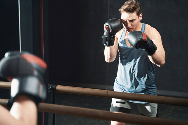 Serious concentrated young male athlete in boxing gloves standing in fighting stance before mirror - Photo, image
