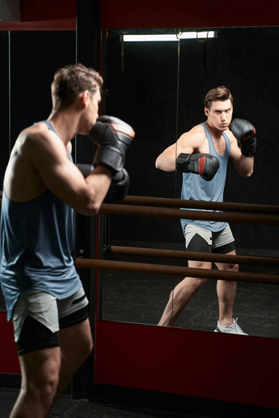 Serious determined strong muscular man in boxing gloves shadow-boxing before gym mirror - Foto, imagen