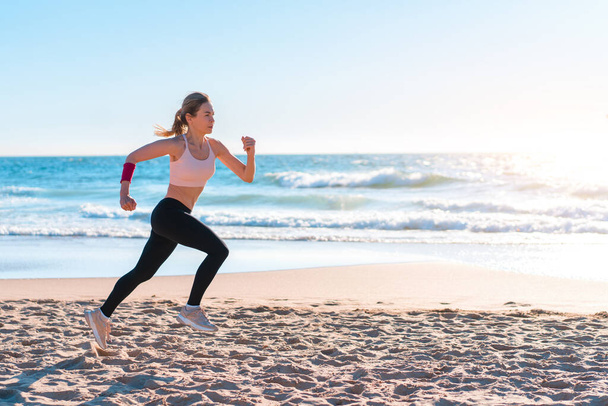 Sporty blonde woman running ocean beach. Young caucasian female exercising outdoors running seashore. Concept of healthy running and outdoors exercise. Active, sporty athlete jogging. Summer active - Φωτογραφία, εικόνα