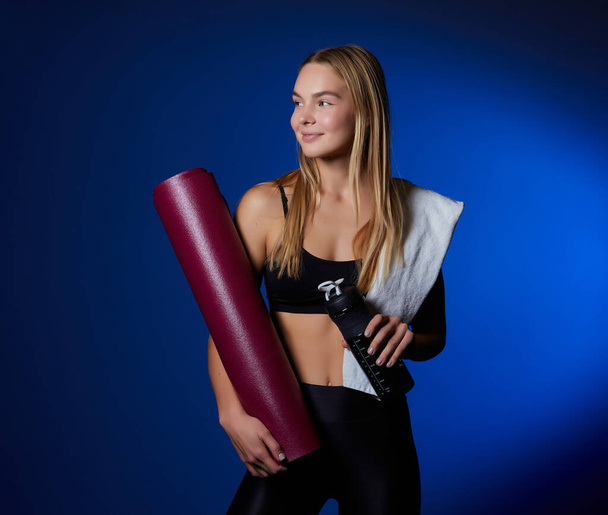 Sports girl, with a towel on her shoulder and a bottle of water in her hands, is resting after training in a dark corner of the gym - Foto, Imagem