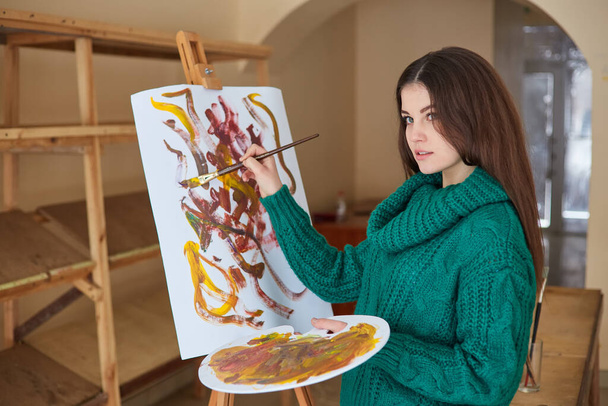 Junge kaukasische Künstlerin, hält Pinsel in der Hand und zeichnet auf Leinwand, arbeitet an einem heimischen Atelier. - Foto, Bild