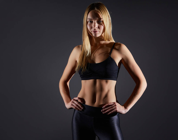 Athletic young blonde girl in sportswear shows muscles on a black background. Great athletic female body - Фото, изображение