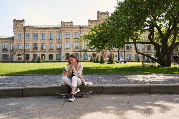 Donna rilassata seduta su asfalto accanto allo skateboard mentre guarda i contenuti sullo schermo del gadget - Foto, immagini